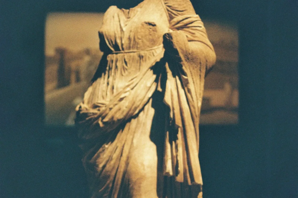 Headless draped marble torso in a museum gallery, arms broken away, lit warm against a dark wall