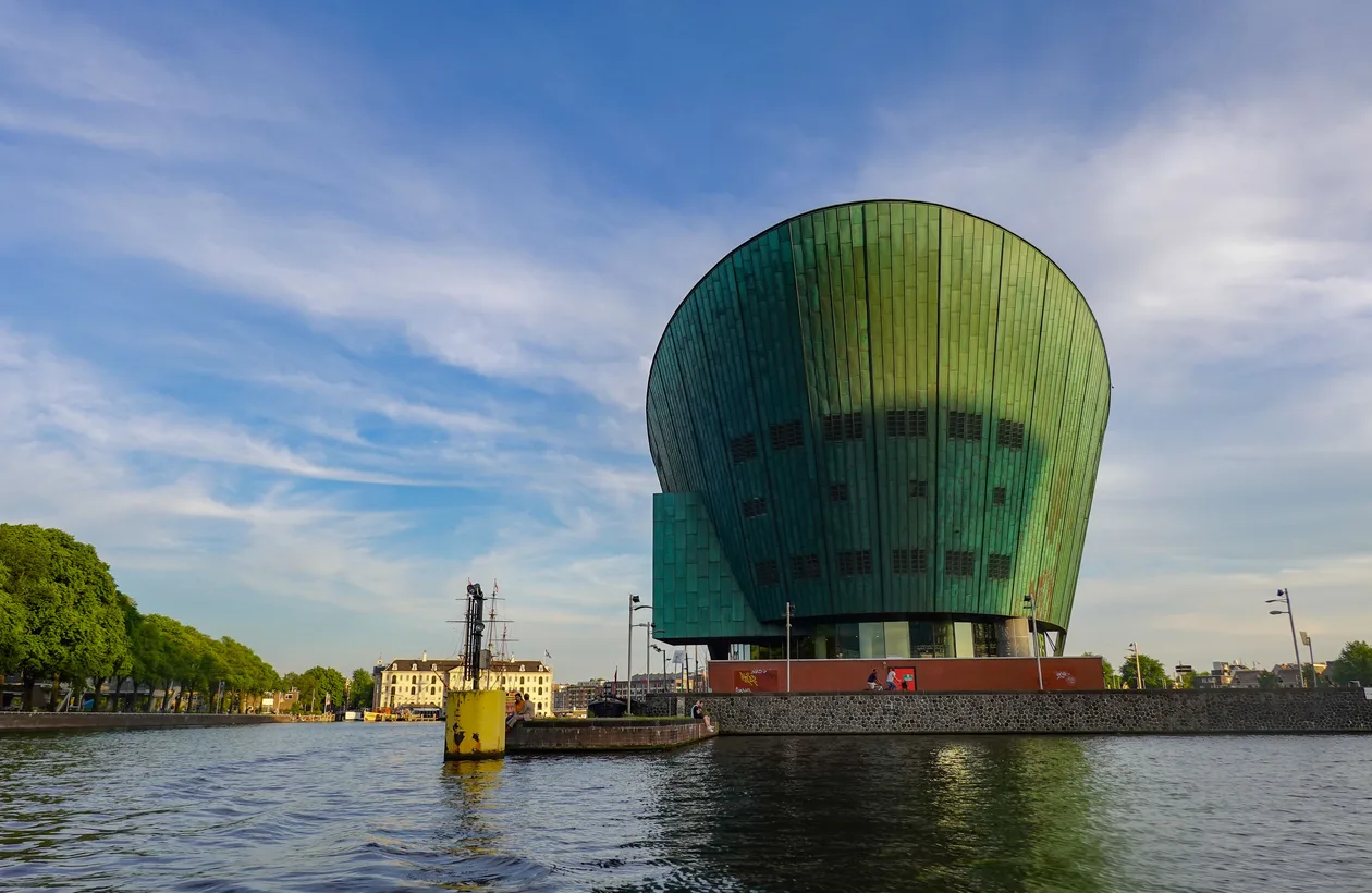 The green copper hull of NEMO Science Museum rising over the Oosterdok water in Amsterdam under a blue sky, with a historic ship and canal houses behind