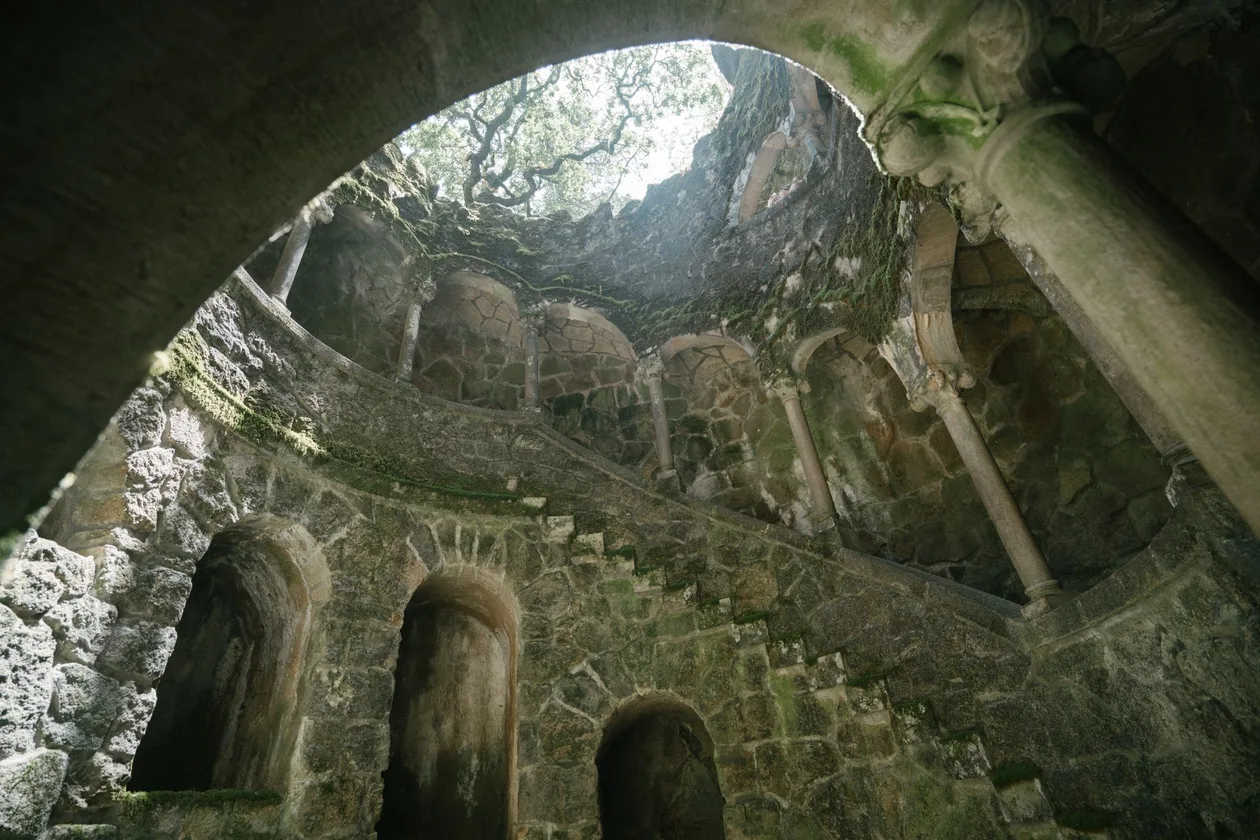 Looking up from inside the Initiation Well at Quinta da Regaleira, mossy stone galleries spiralling up to a circle of trees